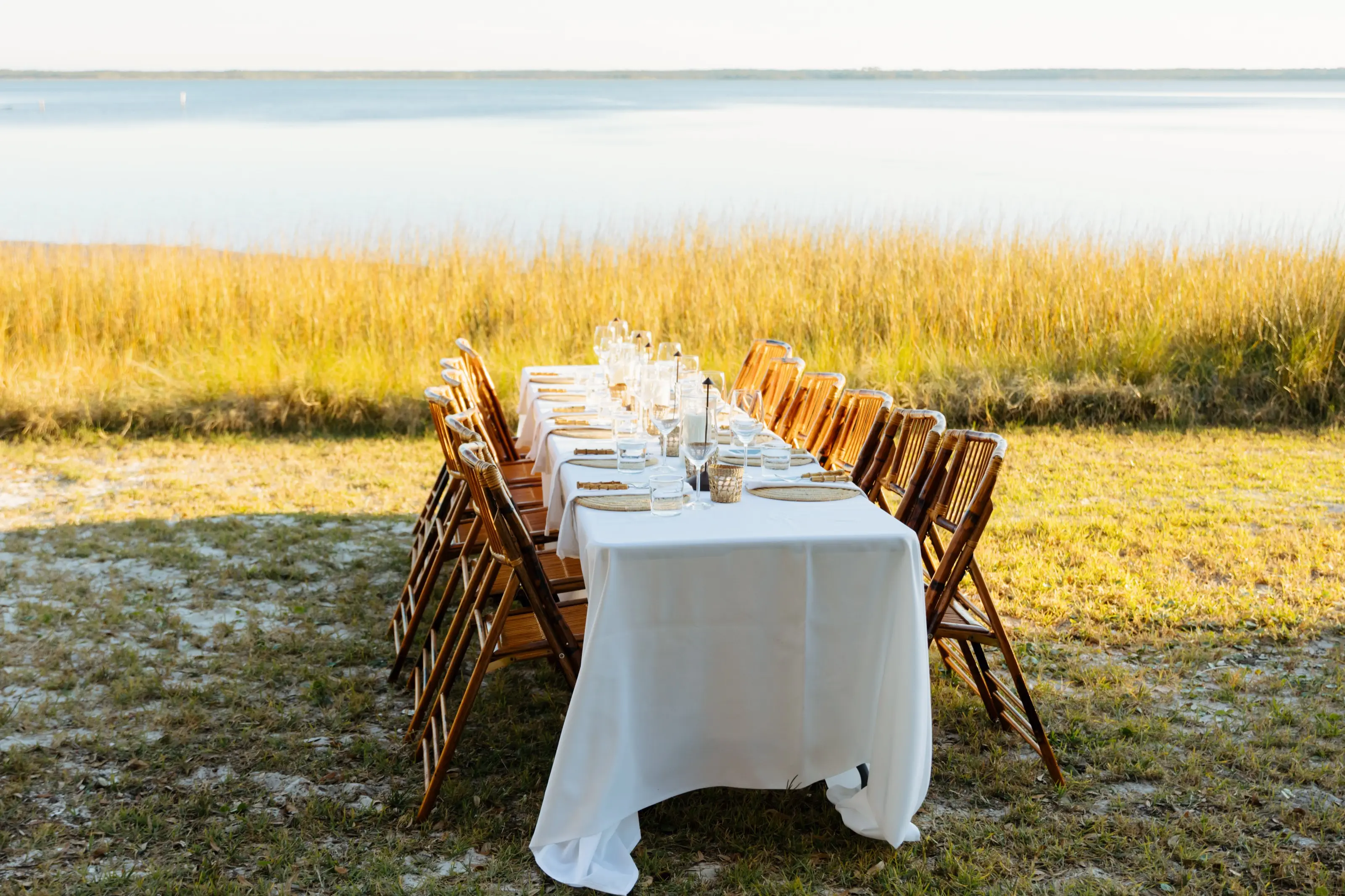 A long table laid in white linen at the edge of the marsh grass, low sun across the water behind it.