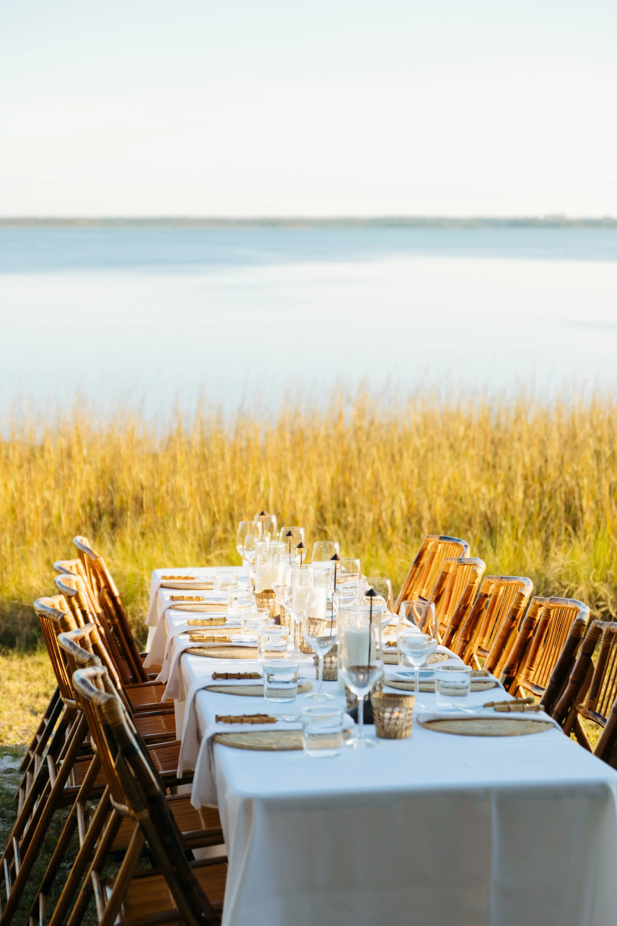 A long table in white linen with bamboo folding chairs, set with glassware and candles at the edge of the marsh grass, the sound beyond.