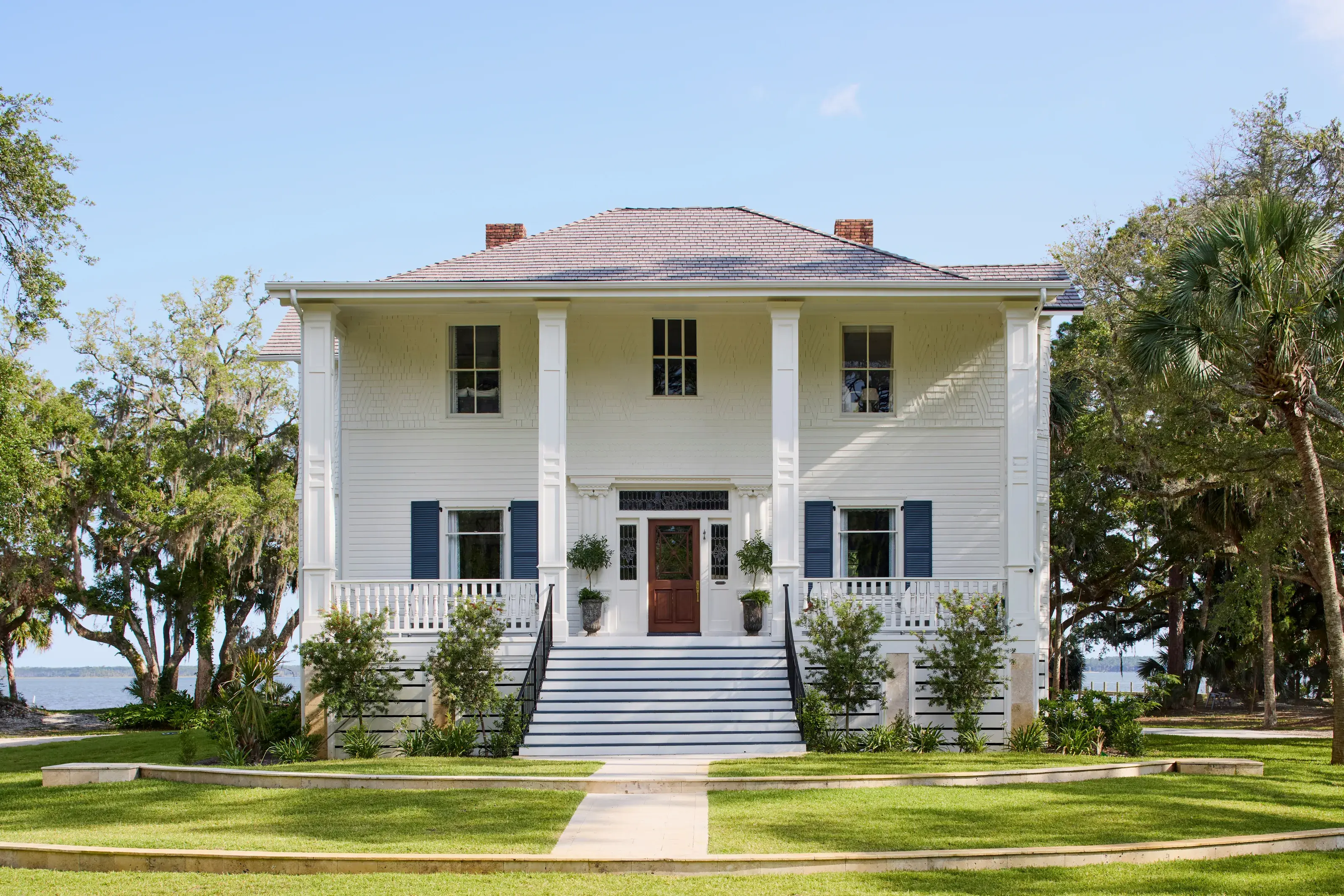 The 1907 Greek Revival house at 11 Mile, white clapboard with blue shutters and a deep two-story porch, framed by live oaks with the water beyond.